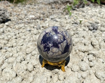May include: A polished, round, blue and gray gemstone sphere on a gold metal stand. The sphere has a swirling pattern of blue and gray.
