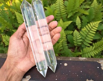 May include: Two clear quartz crystal wands, each with a pointed tip and faceted sides. The crystals are held in a hand, with a backdrop of green ferns and foliage. The crystals are approximately 5 inches long.