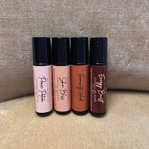 Puede incluir: Cuatro pequeños frascos de vidrio con tapas negras y etiquetas. Las etiquetas son blancas con texto negro y enumeran los nombres de las mezclas de aceites esenciales: "Energy Boost", "Immunity Aid", "Skin Bliss" y "Peace Potion".