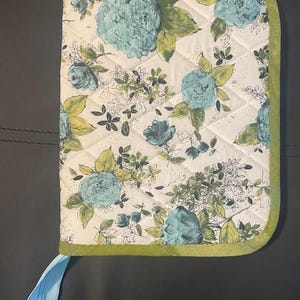 May include: Quilted floral book cover featuring a pattern of blue flowers and green leaves on a cream background. The cover has a green border and a light blue ribbon with a floral design. The cover is designed to protect books.