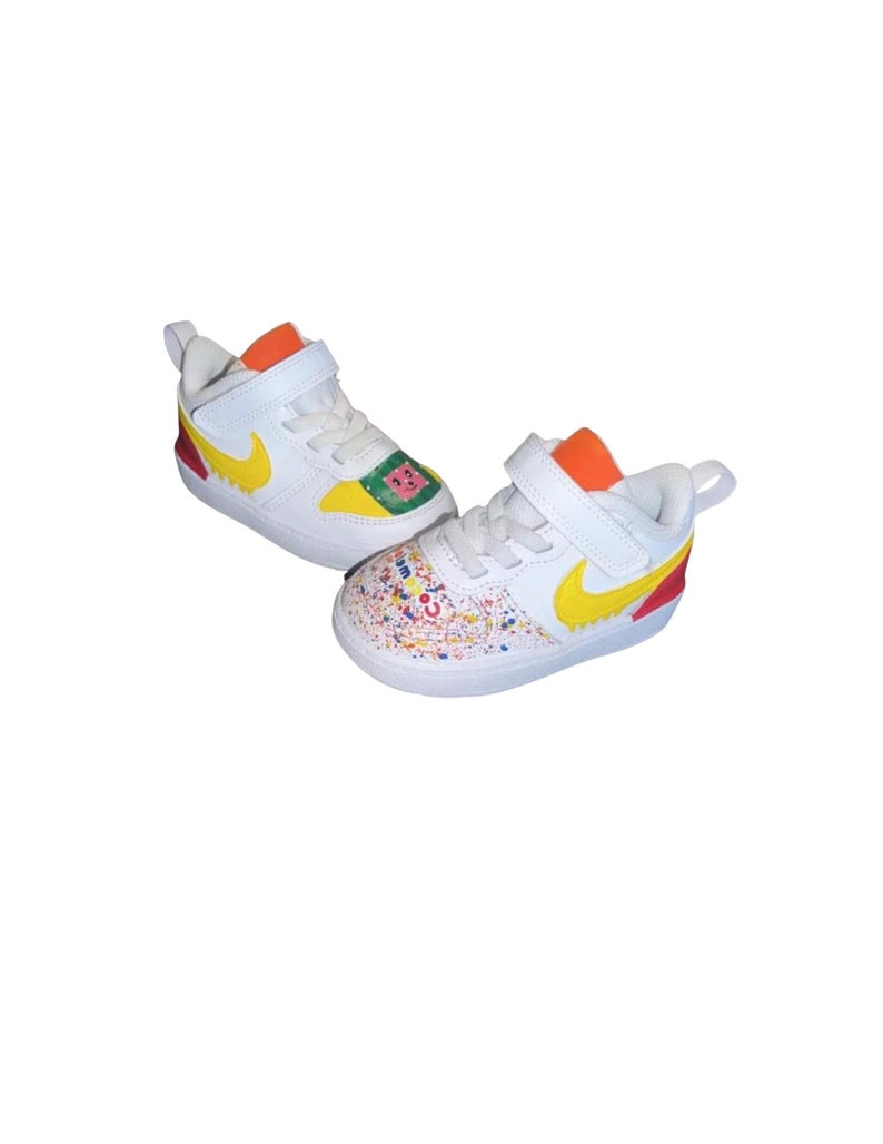 May include: White and yellow Nike sneakers for toddlers. The shoes feature a yellow Nike swoosh, orange accents, and colorful paint splatter details. One shoe has a watermelon graphic.