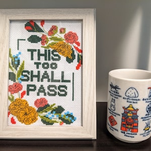 Może przedstawiać: Biały haftowany krzyżyk w ramce z kwiatowym obramowaniem i tekstem "This Too Shall Pass". Obok haftowanego krzyżyka w ramce znajduje się biały ceramiczny kubek z japońskimi znakami i ilustracjami.