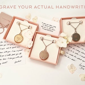 May include: Three open boxes with pink interiors, each containing a gold or silver necklace with a round pendant. The pendants are engraved with handwritten messages. The boxes are surrounded by white flowers.