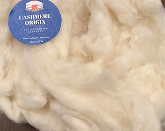 May include: A close-up of a pile of white cashmere wool. The wool is soft and fluffy, and it is labeled as "Cashmere Origin 100% Mongolian Cashmere".