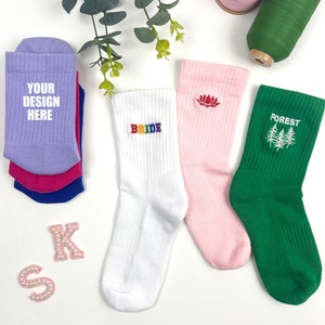 May include: Four pairs of white crew socks with different embroidered designs. One pair says "BRIDE" in rainbow letters, one pair has a pink lotus flower, one pair has a green forest scene, and one pair says "YOUR DESIGN HERE" in white letters.
