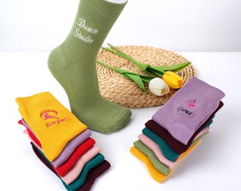 May include: A green ribbed sock with "Dance Studio" embroidered in white. Several stacks of colorful socks are displayed, including yellow, red, and purple, each with a dance-themed design. The socks are on a white surface.