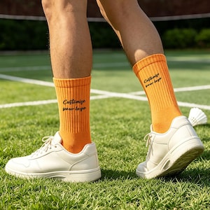 Puede incluir: Un par de calcetines de caña media acanalados de color naranja brillante con el texto negro "Customize your logo" impreso en el lateral de cada calcetín, junto con zapatillas deportivas blancas sobre una pista verde.