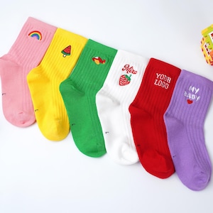 Pode incluir: Seis pares de meias coloridas com desenhos bordados. As meias são rosa, amarelas, verdes, brancas, vermelhas e roxas. Cada meia tem um design diferente, incluindo um arco-íris, uma melancia e o texto "YOUR LOGO".