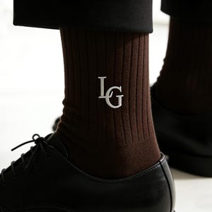 May include: Brown ribbed socks with the embroidered initials "IG" in white. The socks are worn with black dress shoes. The socks are a dark brown color and have a ribbed texture.