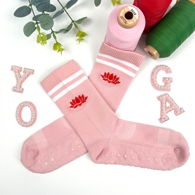 May include: A pair of pink yoga socks with white stripes and a red embroidered lotus flower design. The socks have grippy dots on the bottom.