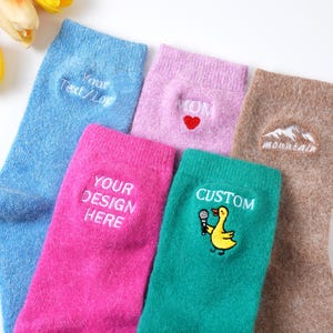 May include: A selection of colourful socks with embroidered designs. The socks are in shades of blue, pink, and brown. Some socks feature text like "Your Text/Logo", "Your Design Here", and "Custom". Other socks have embroidered images of a heart, mountains, and a duck.