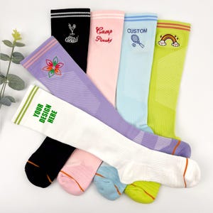 Könnte beinhalten: Sechs Paar Kniestrümpfe mit verschiedenen gestickten Designs. Die Socken sind in verschiedenen Farben, darunter Schwarz, Weiß, Rosa, Hellblau, Lila und Grün. Die Designs beinhalten ein Cocktailglas, die Worte "Camp Pinsky", einen Tennisschläger, einen Regenbogen, eine Blume und die Worte "Ihr Design hier".