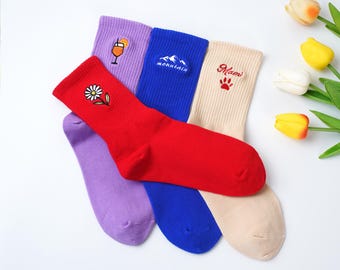 May include: A collection of colorful socks with embroidered designs. The socks are red, purple, blue, and beige. The red sock has a daisy, the purple sock has a cocktail, the blue sock has a mountain, and the beige sock has the word "Mami" and a paw print.