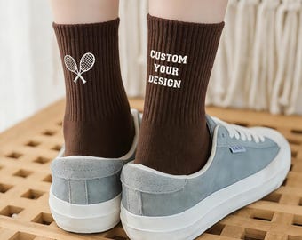 Puede incluir: Calcetines marrones acanalados con el texto blanco "CUSTOM YOUR DESIGN". Un calcetín presenta un gráfico de raqueta de tenis blanca. Los calcetines se usan con zapatillas de deporte azul claro con suelas blancas. Vista desde abajo.