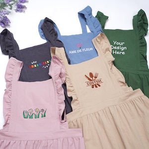 Puede incluir: Una colección de delantales con volantes en varios colores, incluyendo rosa, azul, beige, gris y verde. Cada delantal presenta diseños bordados o texto únicos, como flores, las palabras "EMBROIDERY", "AME DE FLEUR", "CUSTOM" y "Your Design Here".