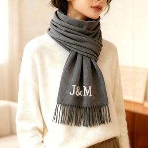 Könnte beinhalten: Grauer Schal mit den eingestickten Initialen "J&M" in Weiß. Der Schal ist um den Hals gewickelt und hat einen Fransenrand. Die Person trägt einen weißen Pullover.