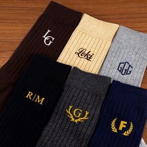 May include: Selection of ribbed socks in shades of brown, black, grey, and navy. Each sock displays embroidered initials or a name in contrasting colours. The socks are presented on a wooden surface.