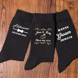 Könnte beinhalten: Drei Paar schwarze Socken mit weißem und goldenem Text. Ein Paar sagt "Atticus GROOMS MAN" mit einer Fliege. Ein anderes Paar sagt "James & Emily March 16th, 2026" mit Eheringen. Das dritte Paar sagt "DANNY Groom 26/04/16".