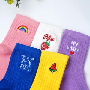 Può includere: Una collezione di calzini a coste colorati. Calzino rosa con un arcobaleno, calzino bianco con una fragola e il nome "Miro", calzino blu con un bulldog e la parola "CUSTOM", calzino giallo con un'anguria e calzino viola con "MY BABY" e un cuore.
