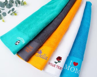 May include: A selection of embroidered hand towels in various colours: teal, grey, orange, white, and blue. Each towel features unique embroidery, including a football, the name "Bella", a rose, and the words "I MOM".