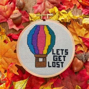 Puede incluir: Un aro de bordado blanco con un globo aerostático colorido y el texto "Let's Get Lost". El aro está rodeado de hojas de otoño.