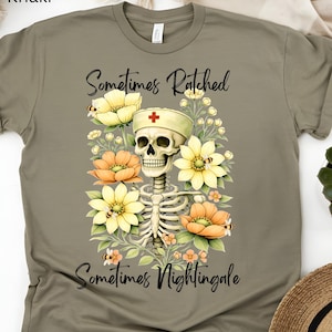 May include: Khaki-colored t-shirt featuring a skeleton wearing a nurse's cap, surrounded by yellow and orange flowers. The text "Sometimes Ratched, Sometimes Nightingale" is written above and below the skeleton graphic.