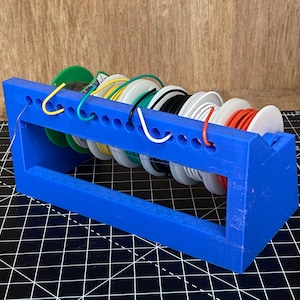Puede incluir: Un soporte de carrete de plástico azul con seis carretes de alambre. Los carretes son de varios colores, incluyendo rojo, blanco, negro, verde, amarillo y azul.