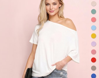 Op de afbeelding: Een witte off-the-shoulder top met korte mouwen. De top wordt gecombineerd met een spijkerbroek. Een zwarte clutch wordt in de linkerhand gehouden. De achtergrond is effen lichtroze. Een kleurenpalet met verschillende kleuren staat rechts.