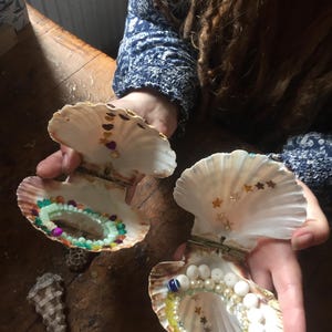 May include: Two open scallop shells functioning as hinged containers. The interior of each shell features a small circular mirror surrounded by colorful beads and pearls. The upper interior of the shells are decorated with small metallic star and heart shapes.