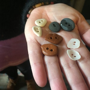 Può includere: Assortimento di sei bottoni unici in varie forme e colori, tra cui marrone, bianco e grigio. I bottoni hanno due fori ciascuno.