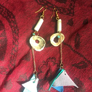 May include: A pair of handmade earrings with gold-coloured hooks. Each earring features a white, cylindrical bead, a shell, and a layered triangle design in shades of blue and grey. The earrings hang from a gold chain.