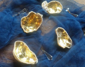 Puede incluir: Varias conchas de ostra decorativas con interior dorado y borde de pequeñas perlas blancas. Las conchas están dispuestas sobre un fondo de tela azul, con algunos elementos decorativos de cristal.