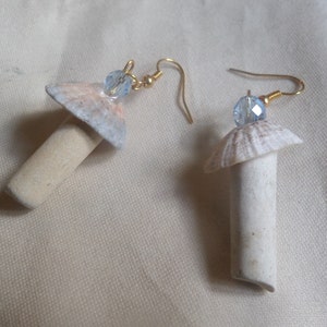 May include: Handmade mushroom earrings. Each earring has a beige, cylindrical stem, a seashell cap with pink and grey hues, and a blue crystal bead. The earrings hang from gold-coloured ear wires. The earrings are a unique accessory.
