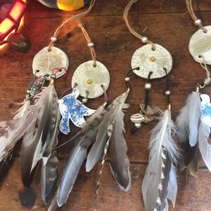 Può includere: Acchiappasogni fatti a mano con dischi in ceramica bianca, adornati con piume grigie, marroni e bianche. Ogni acchiappasogni presenta un cordino di iuta per appenderlo e perline decorative. Gli acchiappasogni hanno uno stile bohémien.
