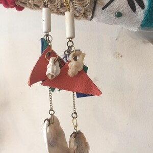 May include: Handmade earrings with a distinctive design. Each earring has a gold-tone hook, a white cylindrical bead, and a red leather triangle. Small seashells and chain accents hang below, with larger shell pendants at the base.