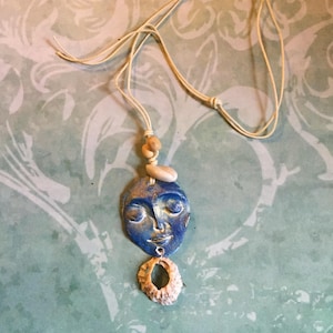 May include: A handcrafted necklace with a blue face pendant, painted with gold accents, and closed eyes. The pendant is suspended from a cream cord, with a seashell and wooden beads. A seashell hangs below the face pendant.