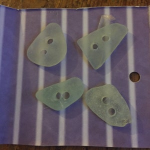 May include: Four sea glass buttons with two holes each. The buttons are a light green color and have a translucent appearance. They are arranged on a purple and white striped background.
