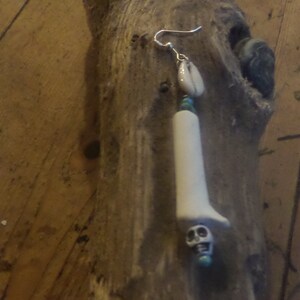 May include: An unusual earring with a white, elongated pendant and a skull at the base. It has a small shell, turquoise beads, and a silver hook. The design is set against a rustic wooden background.