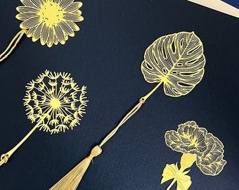 Könnte beinhalten: Vier goldfarbene Lesezeichen aus Metall mit filigranen Ausschnitten in Form eines Gänseblümchens, einer Pusteblume, eines Monstera-Blattes und einer Nelke. Jedes Lesezeichen hat eine passende goldfarbene Kordelquaste am unteren Ende.