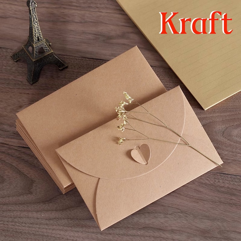 May include: Kraft paper envelopes with a heart-shaped cutout and a sprig of dried flowers. The envelopes are stacked on a wooden surface, with a miniature Eiffel Tower in the background. The word "Kraft" is visible in the upper right corner.
