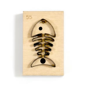 May include: A wooden die-cut featuring a metal fish skeleton design. The die-cut is mounted on a rectangular wooden block. The number "55" is printed in the upper left corner. The fish skeleton is black and has two black dots.
