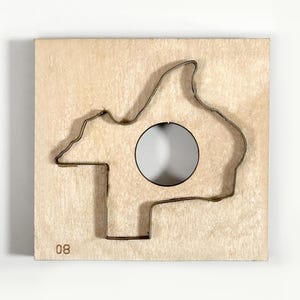 May include: A wooden puzzle piece with a metal outline of a mountain range and a circular cutout. The number "08" is printed in the lower left corner. The wood has a light, natural color.