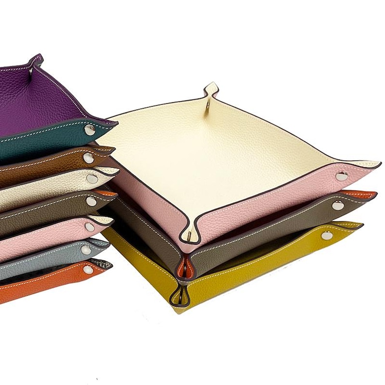 Op de afbeelding: Een stapel lederen trays in verschillende kleuren, waaronder paars, groen, bruin, wit, grijs, roze, blauw, oranje en geel. De trays zijn gemaakt van zacht leer en hebben een gladde, gepolijste afwerking. Elke tray heeft een iets andere maat en vorm, waardoor een visueel aantrekkelijke en unieke set ontstaat.