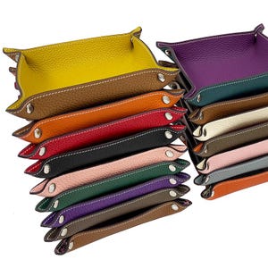 Op de afbeelding: Een stapel van 15 leren trays in verschillende kleuren, waaronder geel, oranje, rood, zwart, roze, groen, paars, bruin en grijs. De trays zijn allemaal even groot en hebben dezelfde vorm, en ze zijn op elkaar gestapeld.