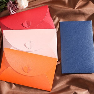 May include: A collection of colorful envelopes in red, pink, and orange, each with a heart-shaped closure. A single blue card sits to the right. The envelopes are arranged on a brown, textured surface, suggesting a stationery or greeting card theme.