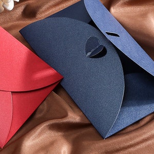 May include: Two decorative envelopes, one red and one navy blue, are displayed on a brown satin surface. The red envelope features a heart-shaped cutout, while the blue envelope has a heart-shaped opening and a slit. The envelopes are likely for invitations or cards.
