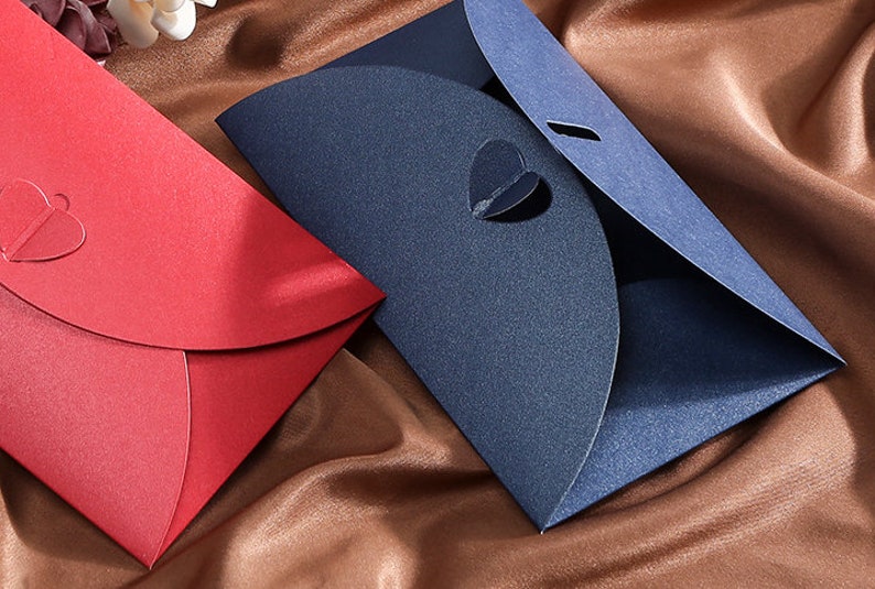 May include: Two decorative envelopes, one red and one navy blue, are displayed on a brown satin surface. The red envelope features a heart-shaped cutout, while the blue envelope has a heart-shaped opening and a slit. The envelopes are likely for invitations or cards.