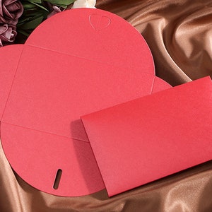 May include: A red, heart-shaped invitation card with a matching envelope. The card is open, revealing its unique shape and a small heart cutout. The envelope is rectangular and sits beside the card on a brown satin surface.