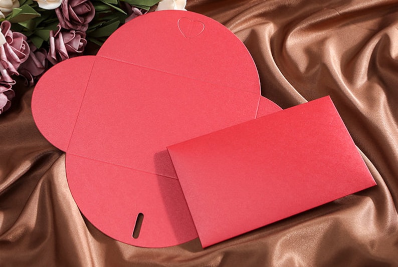 May include: A red, heart-shaped invitation card with a matching envelope. The card is open, revealing its unique shape and a small heart cutout. The envelope is rectangular and sits beside the card on a brown satin surface.