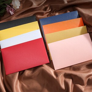 May include: A collection of colorful envelopes in various shades, including red, black, yellow, white, blue, orange, gold, and pink. The envelopes are arranged in stacks, showcasing their different colors and textures. The background is a brown, silky fabric.